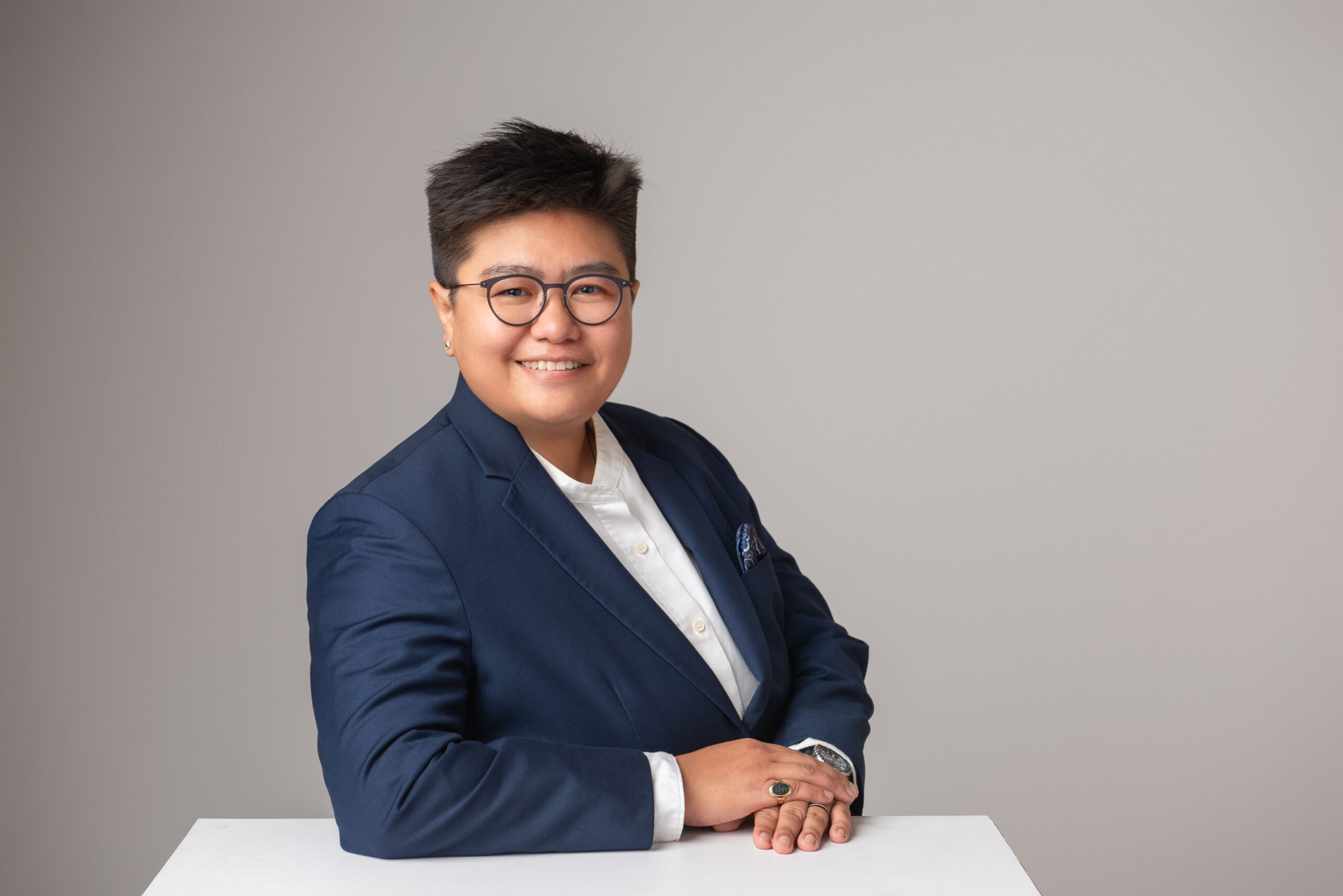 Profile photo of Yu Yu Din, resting on a white table - professionally shot in studio with grayish gradient background. She's wearing a dark blue suit with a white button down shirt with stand collar.